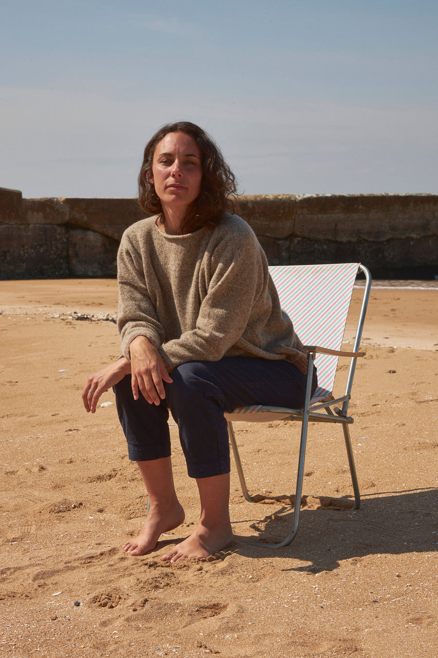Woman wearing an oversized boxy wool jumper, sat on deckchair on Margate beach. Hand knitted jumper, made by British indie brand. Displaying pure 100 percent soft wool knitwear for best capsule winter wardrobe. Baggy fit, in marl brown and natural merino lambswool. Affordable sustainable knitwear made by ethical brand Rove knitwear.