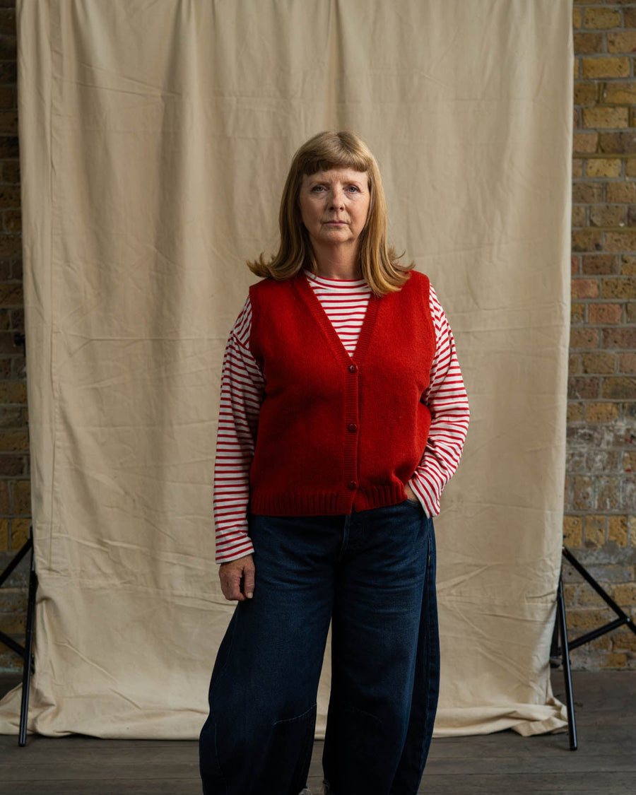 Person wearing a red vest over a striped shirt with blue jeans, standing against a beige backdrop.