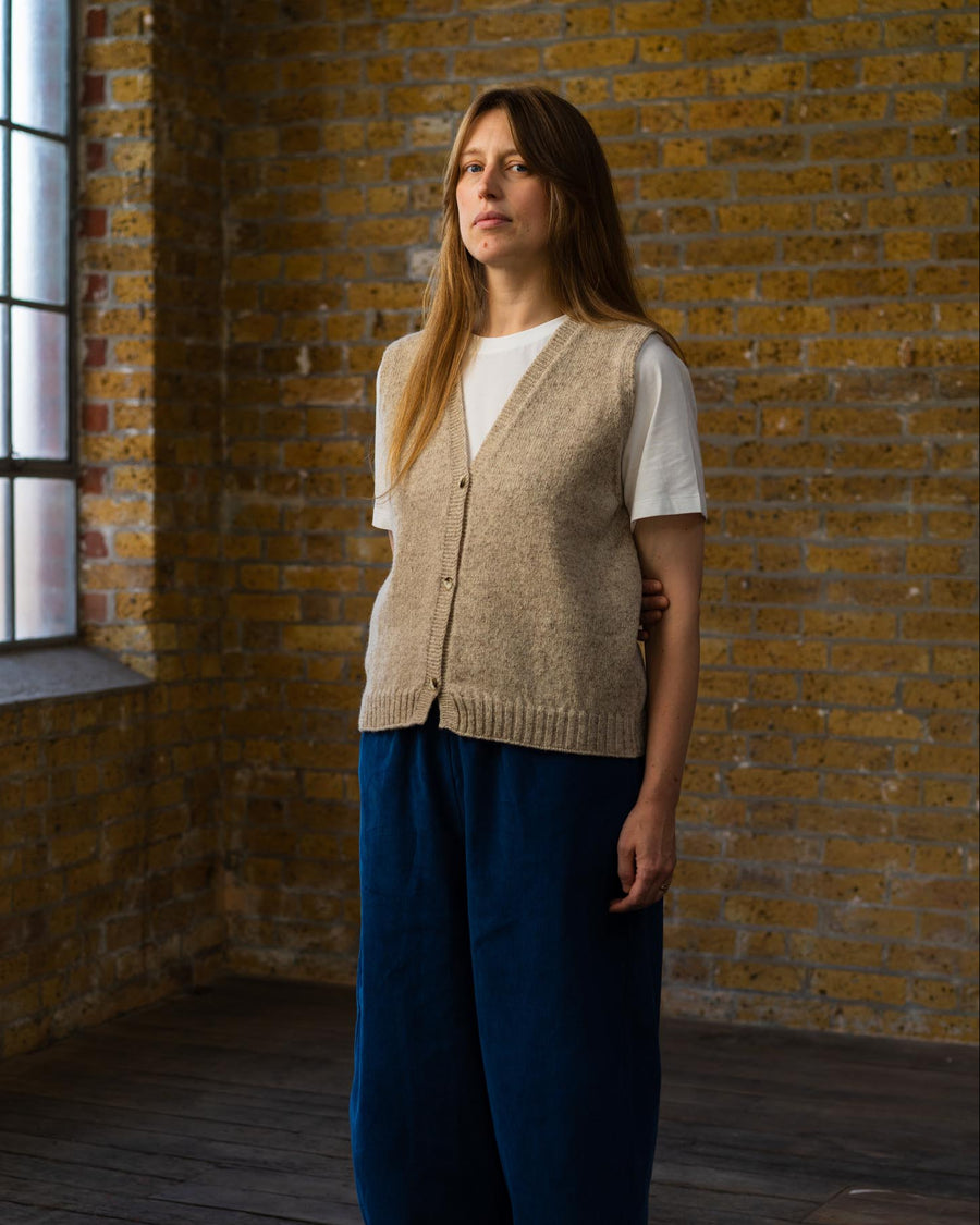 Woman wearing a beige vest and blue pants standing in a room with brick walls and wooden floor.