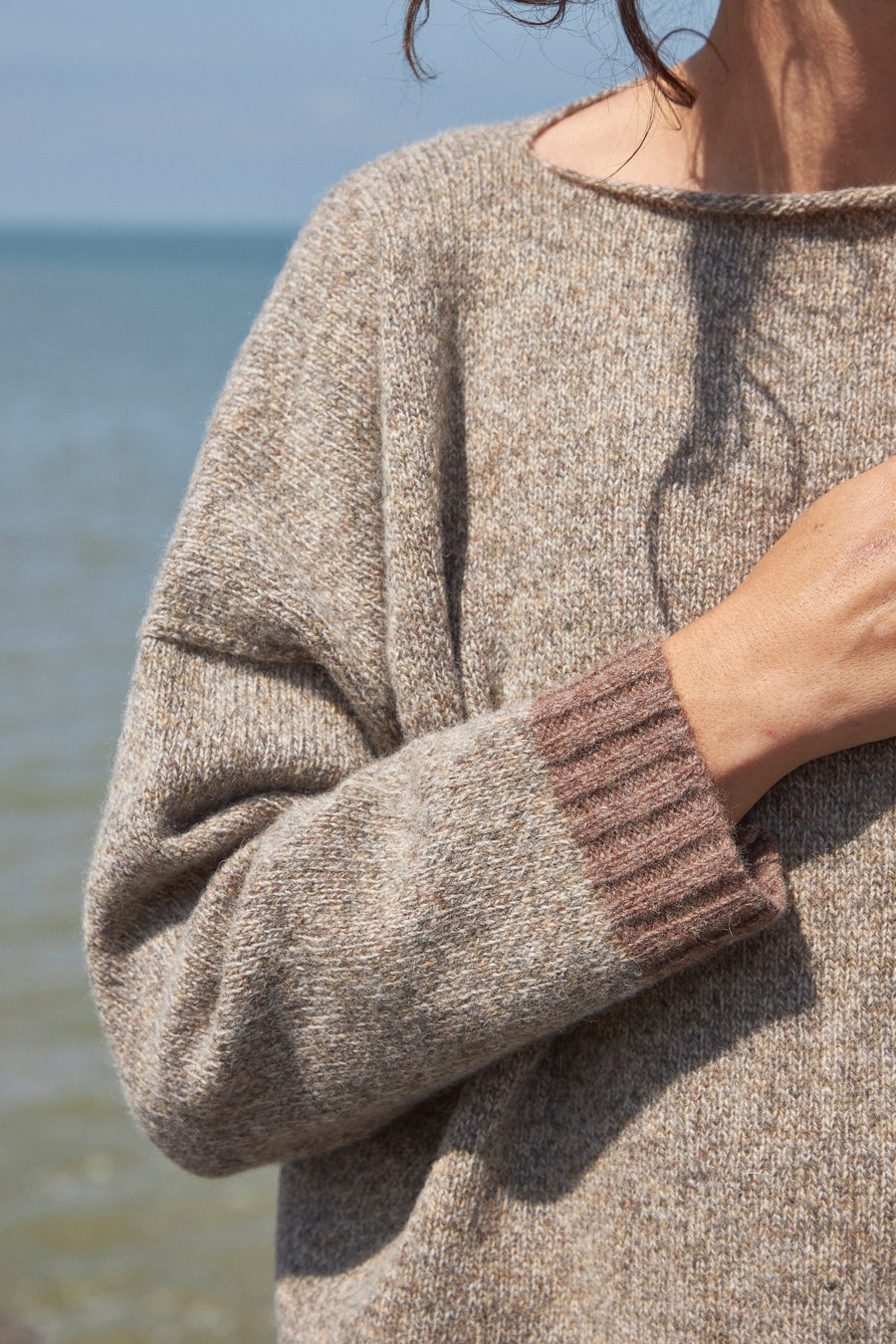 Woman wearing an oversized boxy wool jumper, stood by the seaside in Margate. Hand knitted jumper, made by British indie brand. Displaying pure 100 percent soft wool knitwear for best capsule winter wardrobe. Baggy fit, in marl brown and natural merino lambswool. Affordable sustainable knitwear made by ethical brand Rove knitwear.